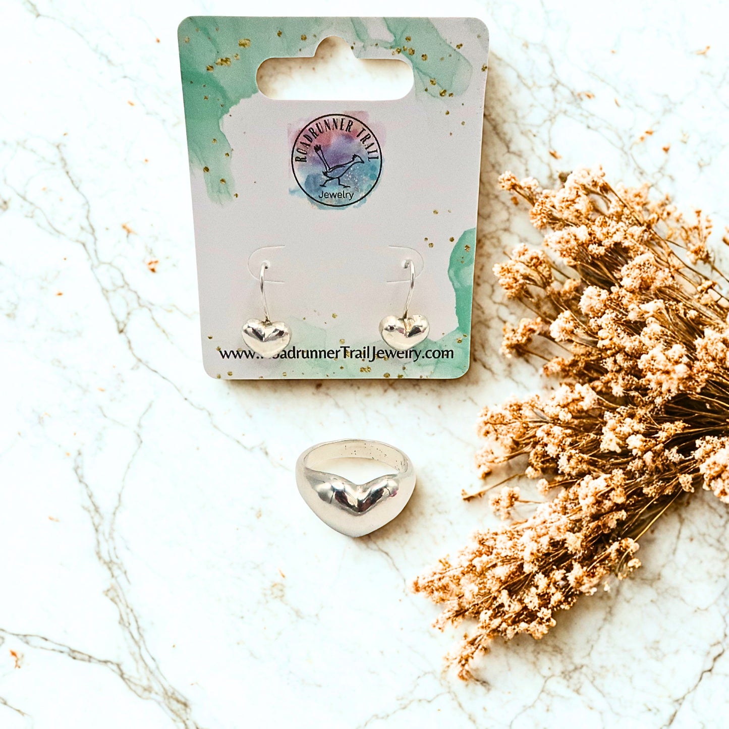 Heart-shaped earrings and puffy heart sterling silver ring on a branded card with dried flowers on a marble surface
